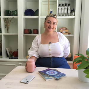 A photo of a person sitting at a cafe table, holding a pink smoothie, and smiling.