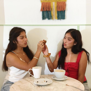 A photo of two identical twins at a coffee shop, smiling at each other and holding up pieces of a scone so they touch, as if saying cheers.